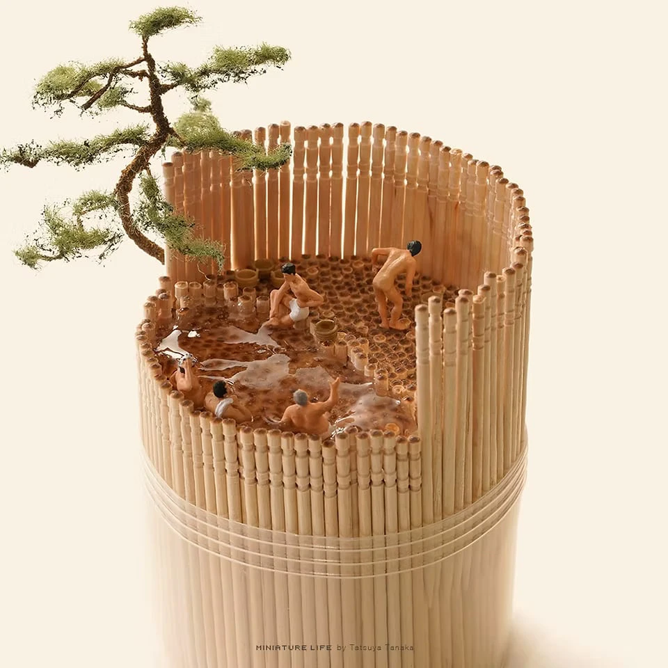 An open-air bath inside a bundle of toothpicks, where tiny figures soak in the water while a miniature pine leans over the fence