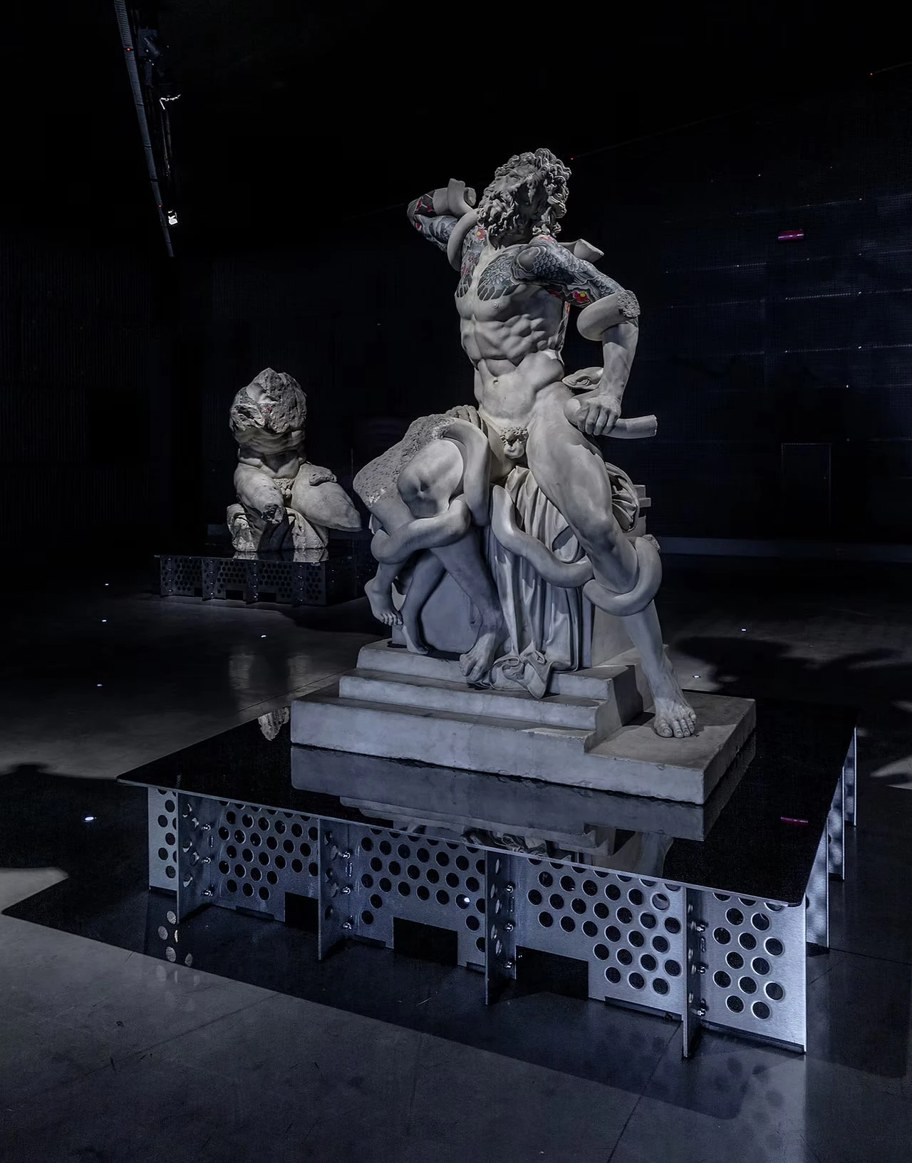 A male figure entwined by serpents on a perforated metal plinth in a dark gallery, tattoos on his shoulders and arms, another sculpture behind