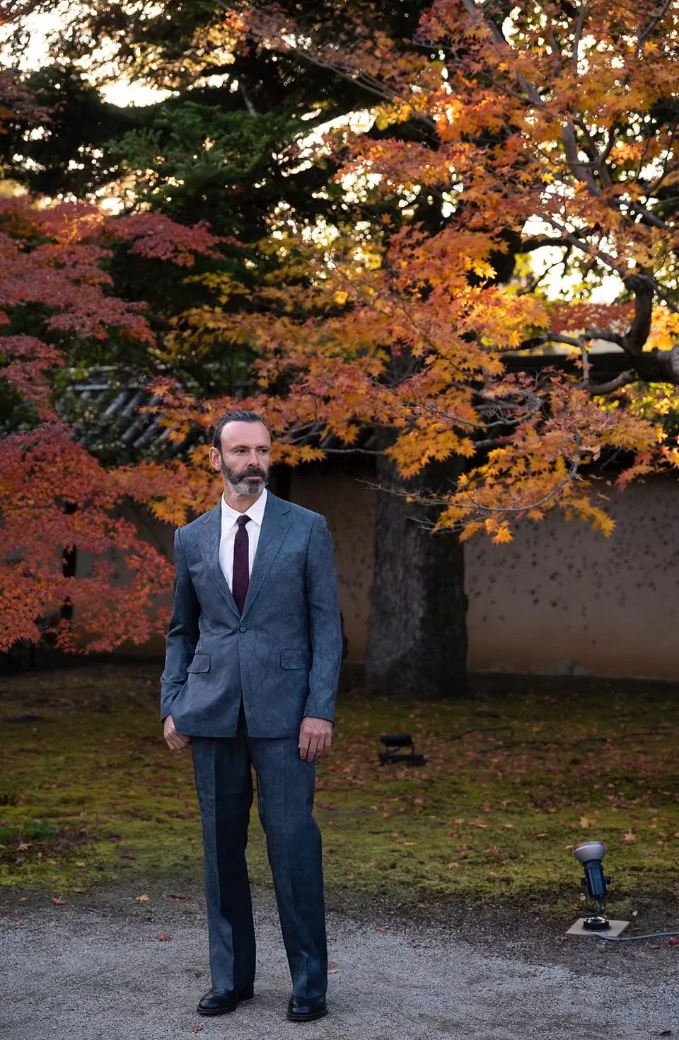 A man in a finely branch-patterned, grey-blue suit standing before an earthen wall beneath autumn maples