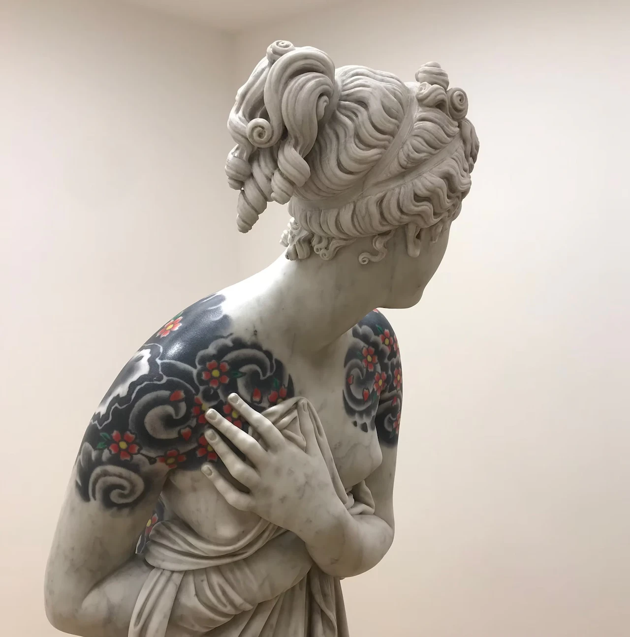 The upper body of a female figure against a cream wall, a hand resting on a shoulder covered in black waves and red cherry blossoms