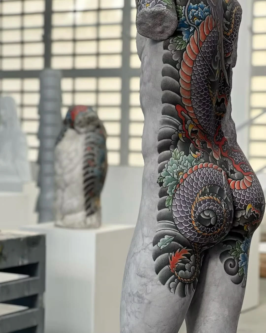 Side view of a torso's hip tattooed with a dragon and peonies, in a bright room with another tattooed sculpture behind