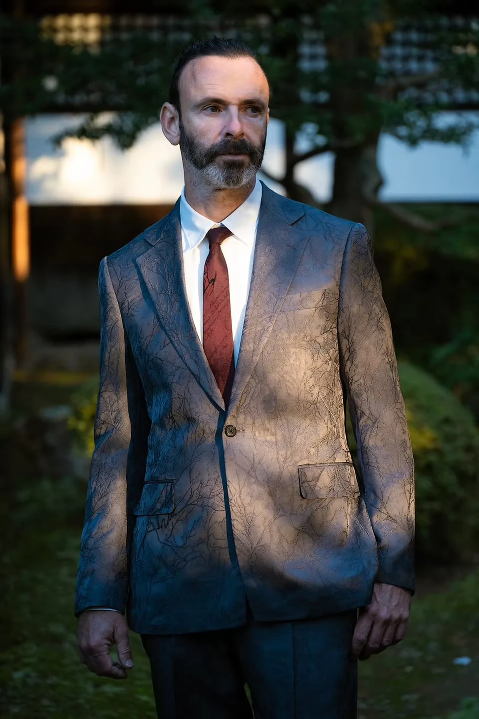 A man in a jacket with a branch-like pattern, white shirt and red tie, raking light falling across the cloth