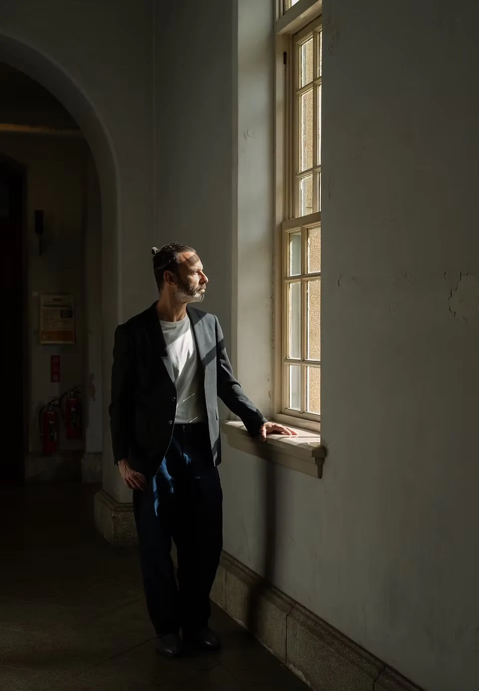 A man in a dim corridor resting a hand on the sill of a tall window, looking out into the light