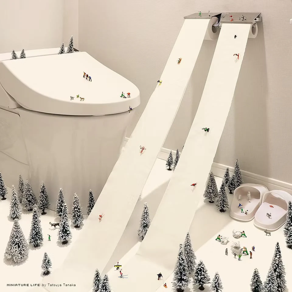 Two lengths of toilet paper hanging from a wall holder become ski slopes, with tiny skiers descending among snow-dusted miniature trees on the bathroom floor