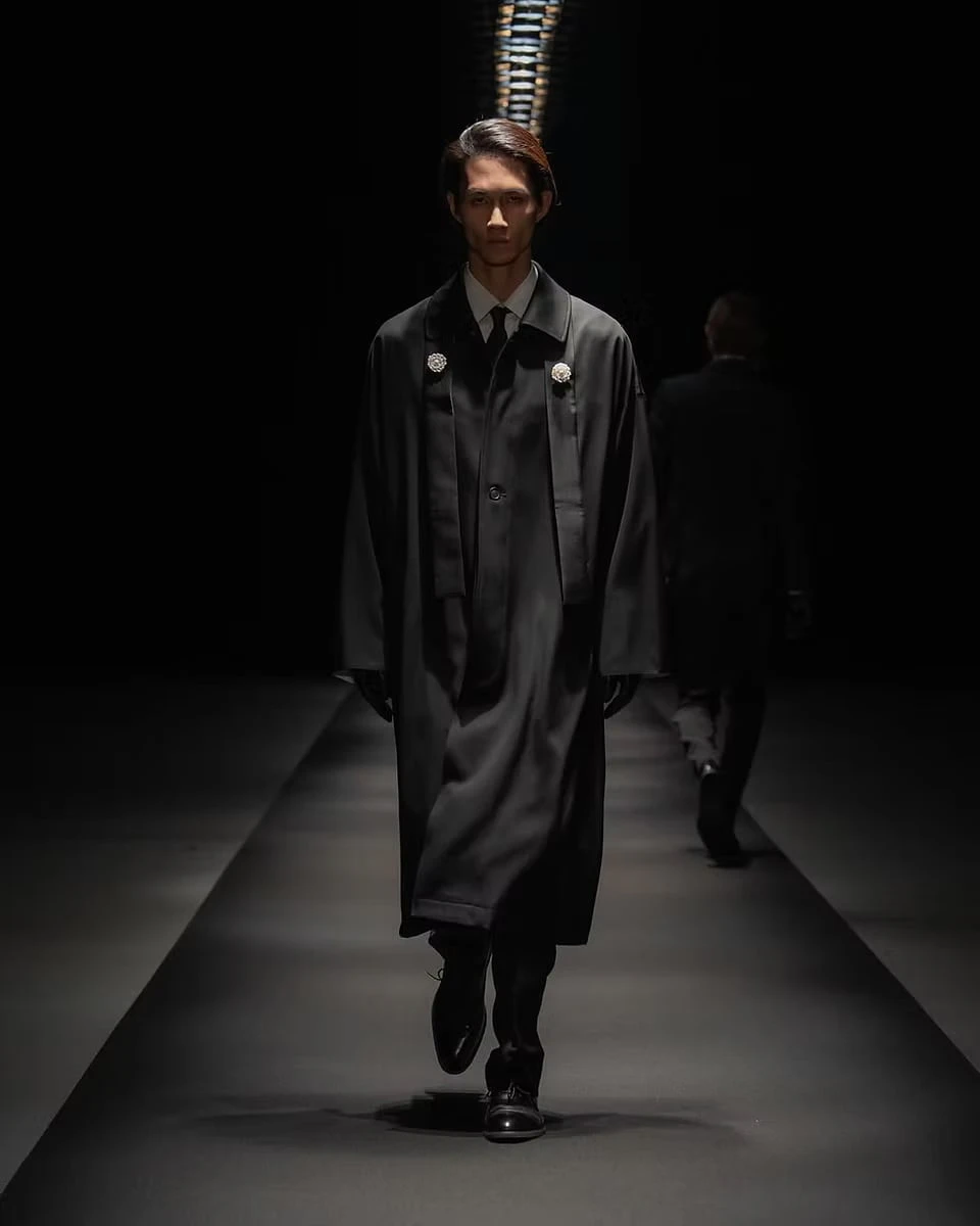 A model on the runway in a long black coat with two brooches pinned at the collar