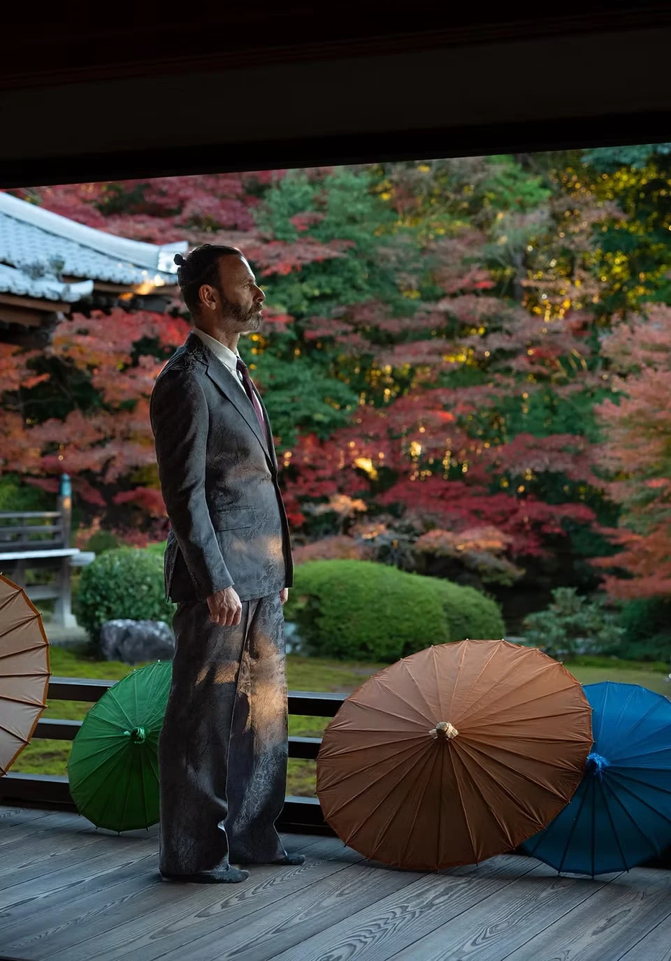 A man in a lustrous patterned suit standing on a temple veranda, coloured paper umbrellas at his feet and an autumn garden beyond
