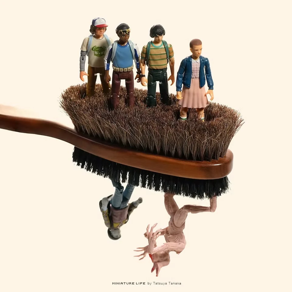 Four child figures stand on the bristles of a clothes brush, while a figure and a creature hang upside down from its underside