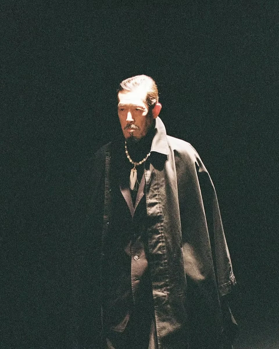 A moustached man lit against darkness, a black cape-like coat over his jacket and a necklace with a white pendant
