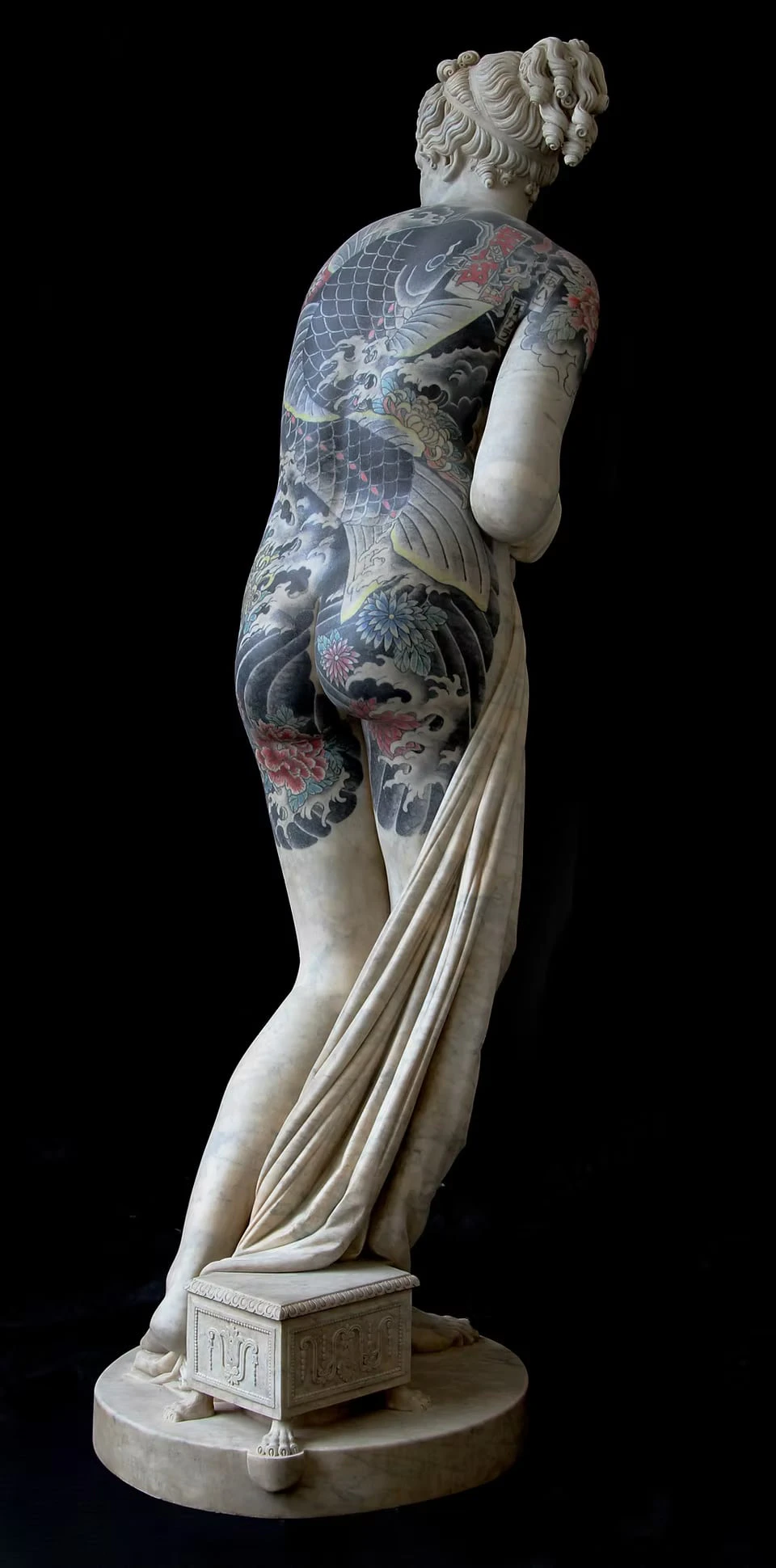 A female figure seen from behind against a black background, her back and legs covered in a tattoo of a carp, waves and peonies