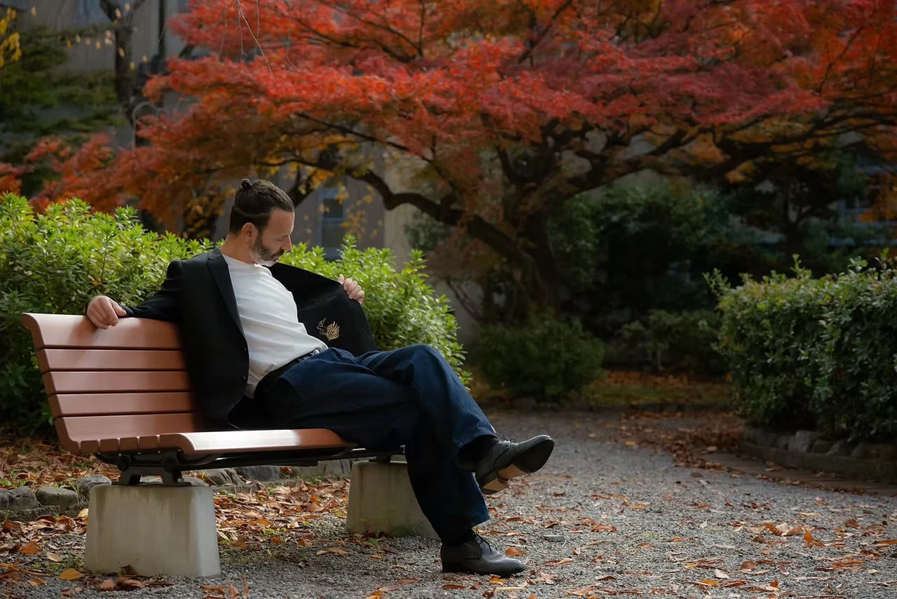 A man seated on a bench beneath a red-leaved maple, glancing into the lining of his black jacket