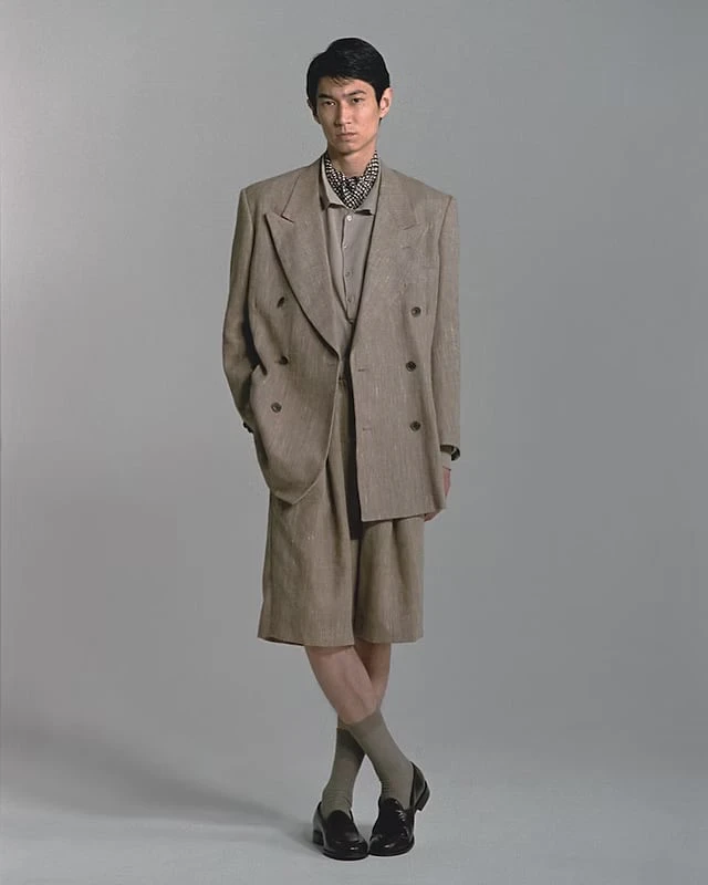 A man in a beige double-breasted jacket with matching shorts, a patterned neckerchief and leather shoes