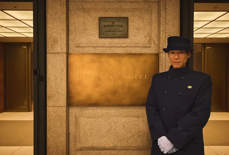 A doorman in a cap and white gloves standing before a stone wall bearing an Imperial Hotel plaque
