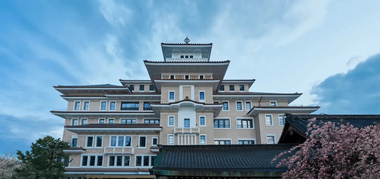 The exterior of the former Yasaka Kaikan under a blue sky, its castle-like tiered tower rising behind cherry blossom