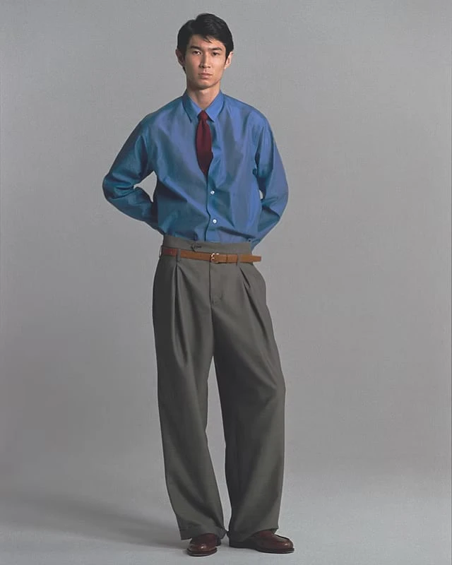 A man in a blue shirt, red tie and belted grey pleated trousers, standing with his hands behind his back