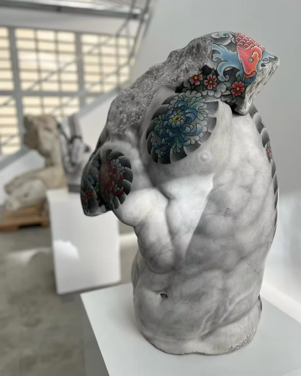A male torso with rough, broken stone surfaces, a red carp and cherry blossoms tattooed on the shoulder and a blue chrysanthemum on the chest