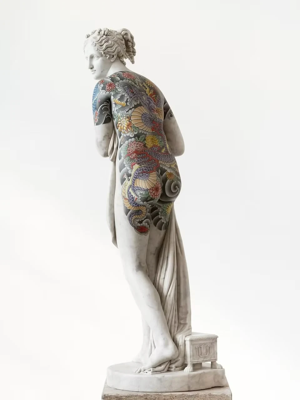A marble female figure seen from behind against a white background, a vivid tattoo of a dragon, chrysanthemums and waves covering her back and hips