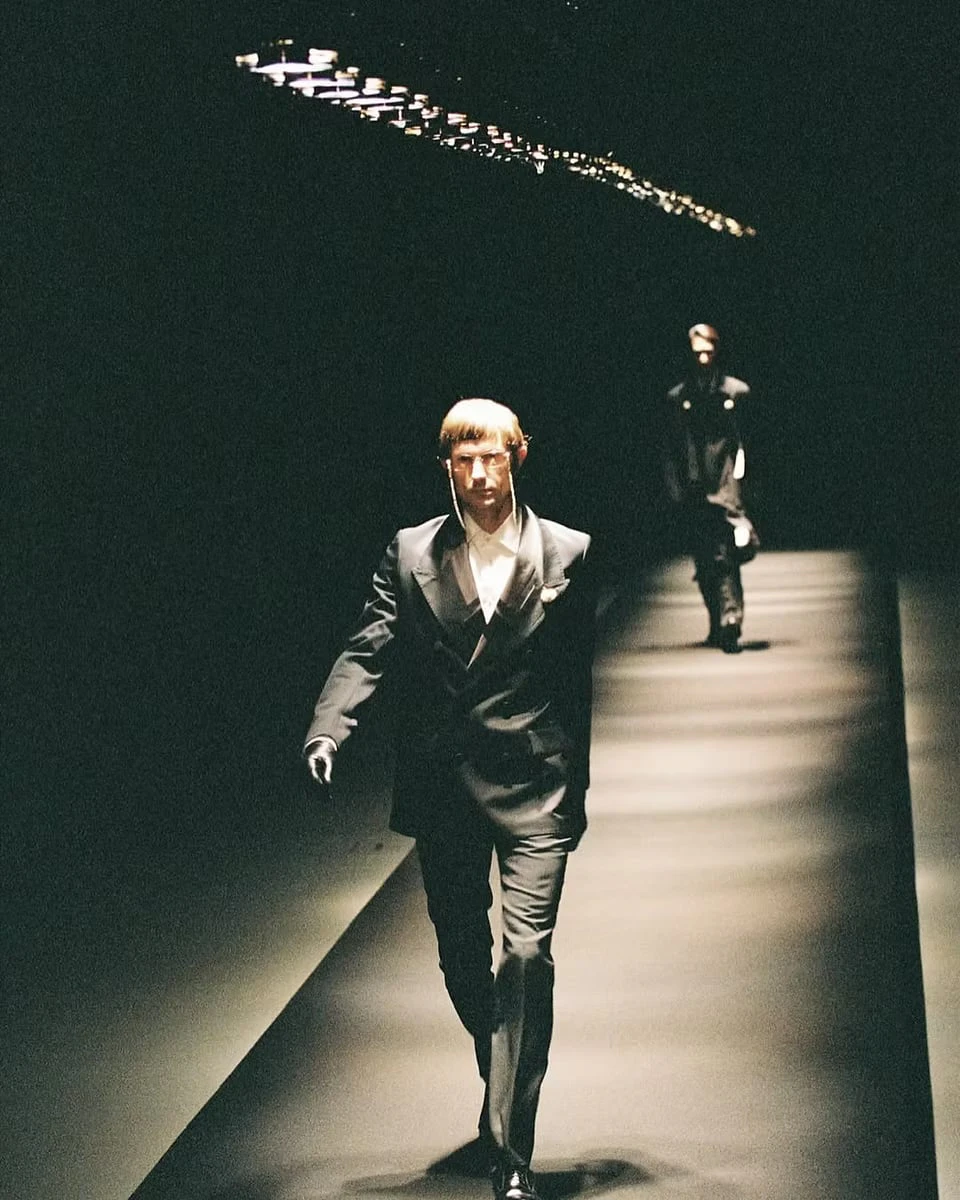 A model walking a dark runway in a black double-breasted suit, white shirt and gloves, with another model following behind