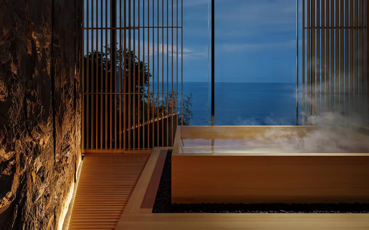 A bathing room framed by a rough rock wall and slender wooden screens, steam rising from a wooden tub before an open view of the sea at dusk