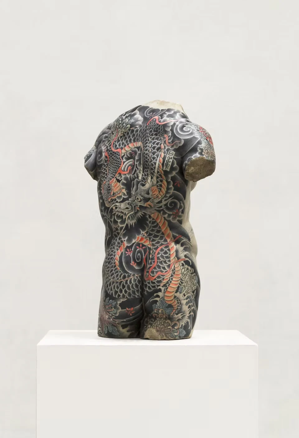 A headless, armless male torso on a white plinth, its back covered by a black and vermilion dragon