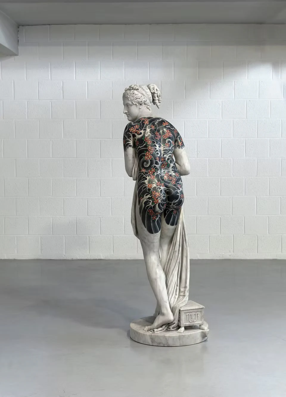 A marble female figure seen from behind in a room with white block walls, black waves and cherry blossoms tattooed from her back to her legs