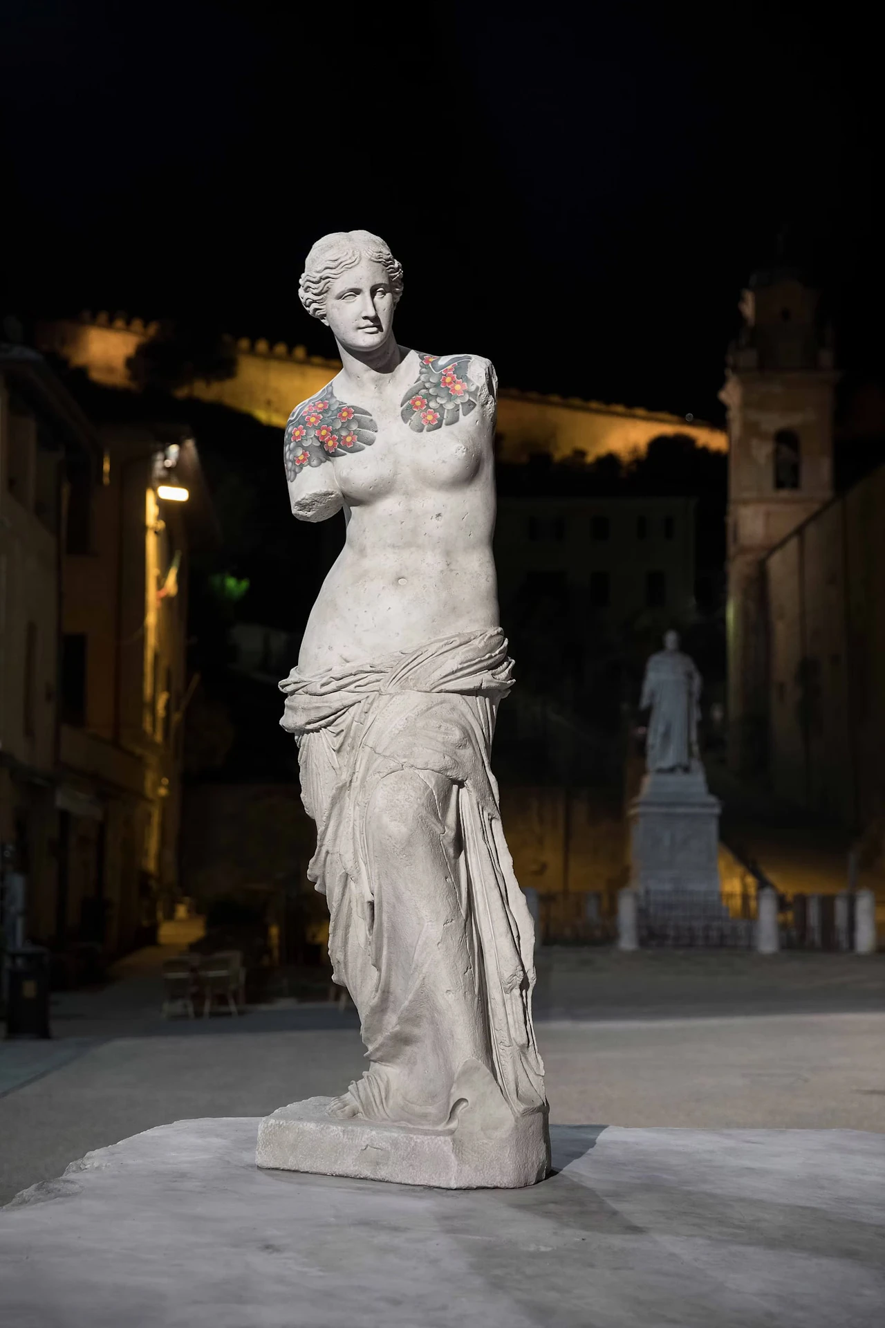 An armless female figure standing on a stone block in a square at night, flowers tattooed on both shoulders, with a statue and bell tower beyond