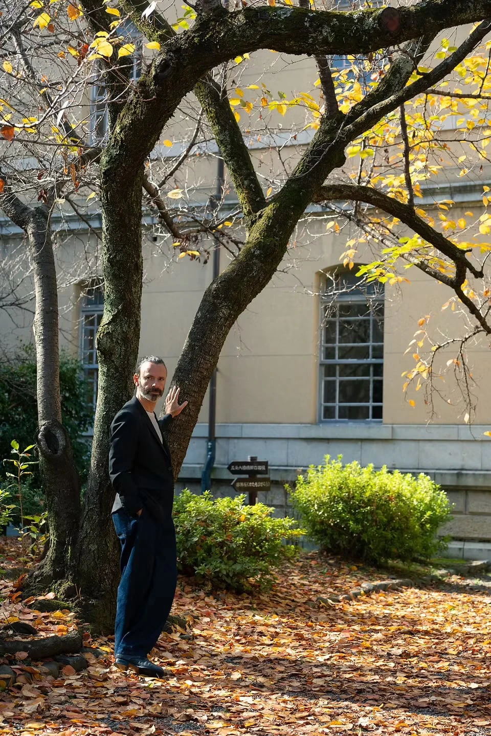 A man in a black jacket resting a hand on a tree trunk in a garden strewn with fallen leaves