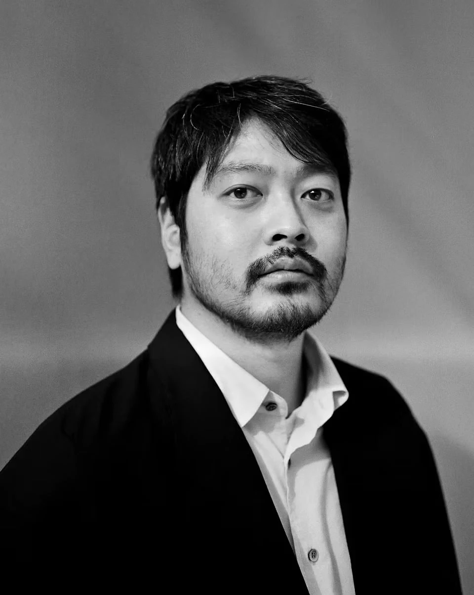 A black-and-white portrait of Soshi Otsuki in a dark jacket and white shirt against a plain backdrop