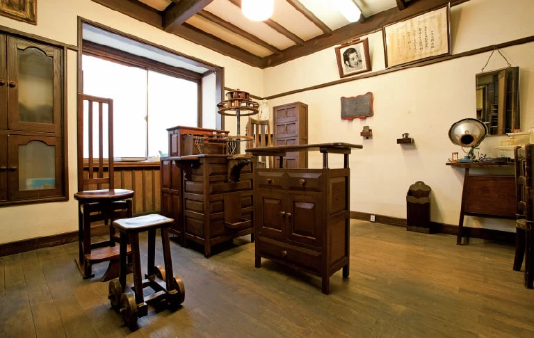 A consulting room with a wooden floor, wooden examination chairs, a medicine cabinet and cupboards, with framed pictures and a portrait on the walls