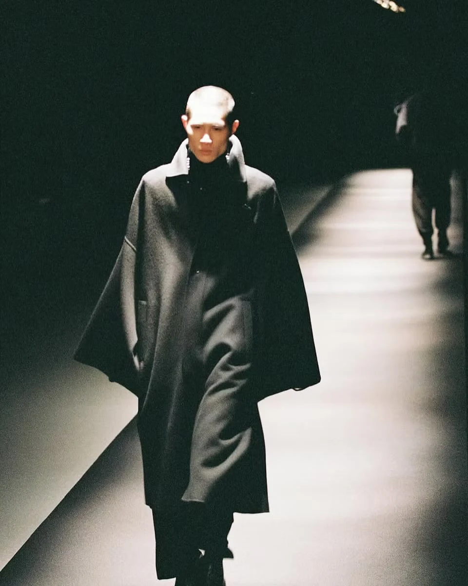 A model with close-cropped hair walking a dim runway, a loose black overcoat swinging with each step