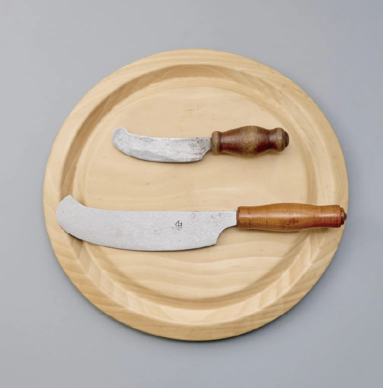 A fruit knife and a bread knife with wooden handles resting on a round plain-wood bread board