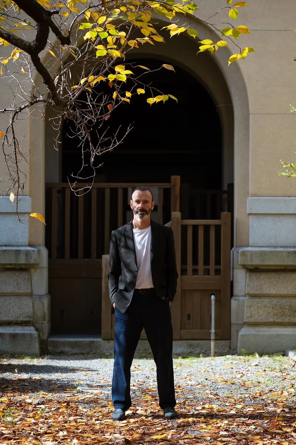 A man in a black jacket standing beneath yellow-leaved branches before an arched entrance