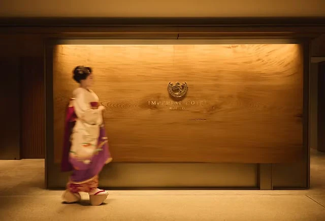 A woman in kimono walking past a wooden wall bearing the Imperial Hotel crest