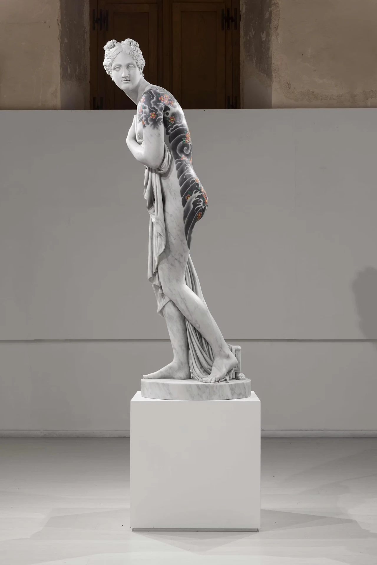 Side view of a female figure on a white plinth, waves and cherry blossoms tattooed across her back and hip