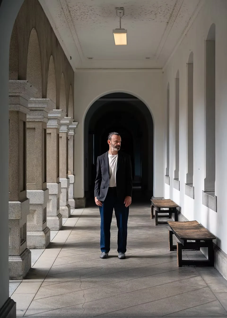 A man in a black jacket standing in a white arcade lined with stone pillars and wooden benches