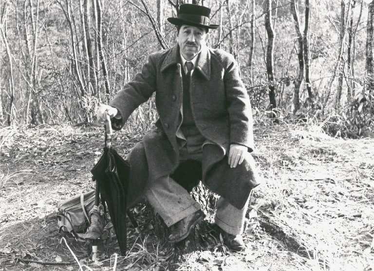 A black-and-white photograph of a man in a hat and overcoat sitting on a tree stump in a woodland, holding an umbrella