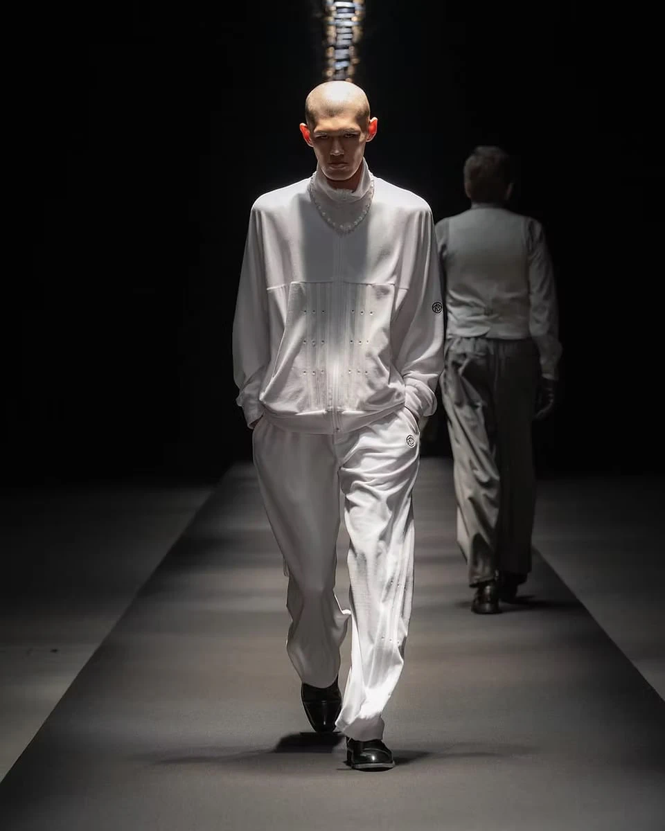 A model on the runway in a white zip-up top and trousers, worn with a string of white beads