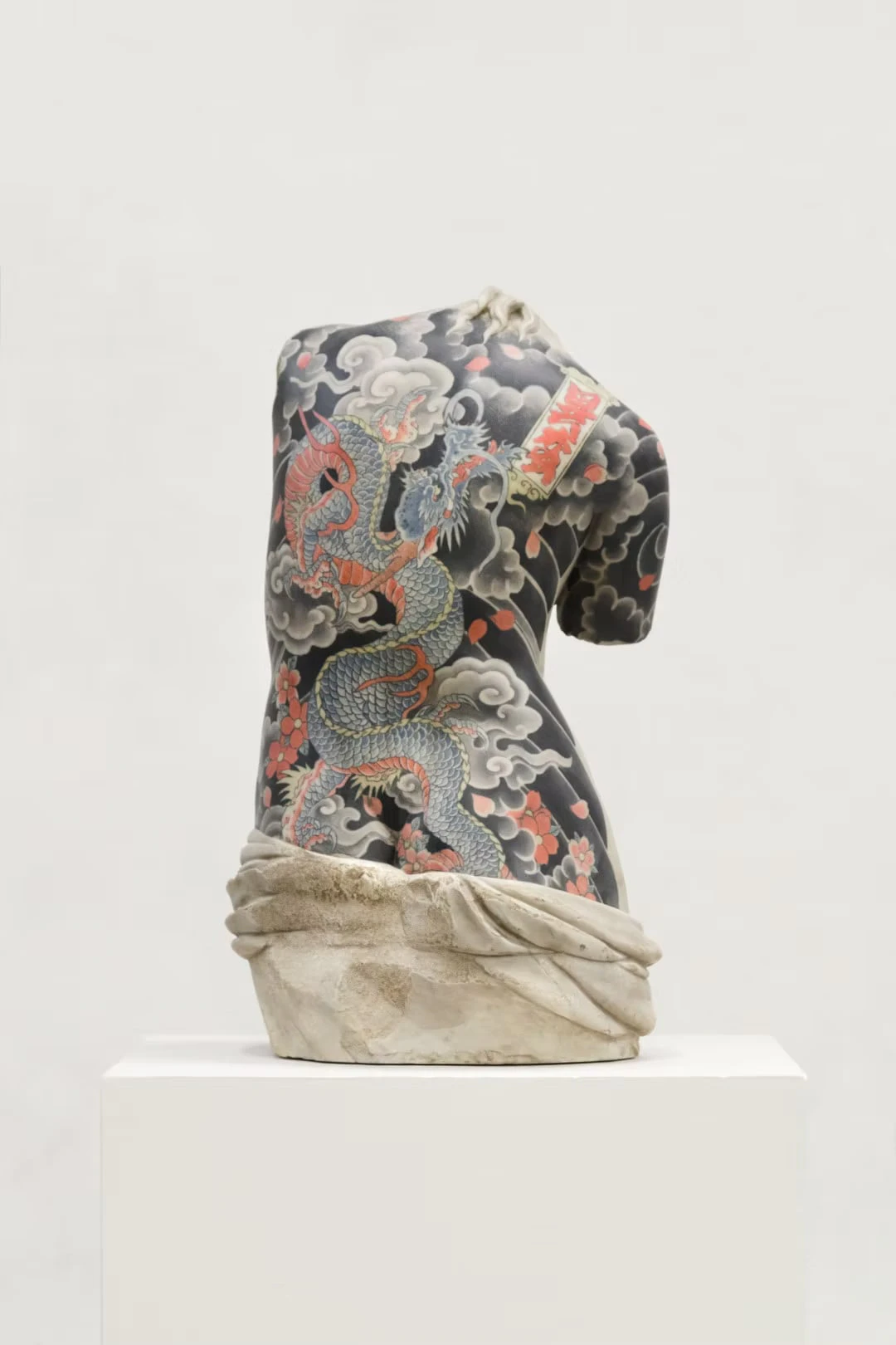 A female torso seen from behind on a white plinth, a dragon among clouds, cherry petals and a red tablet-like motif across the back