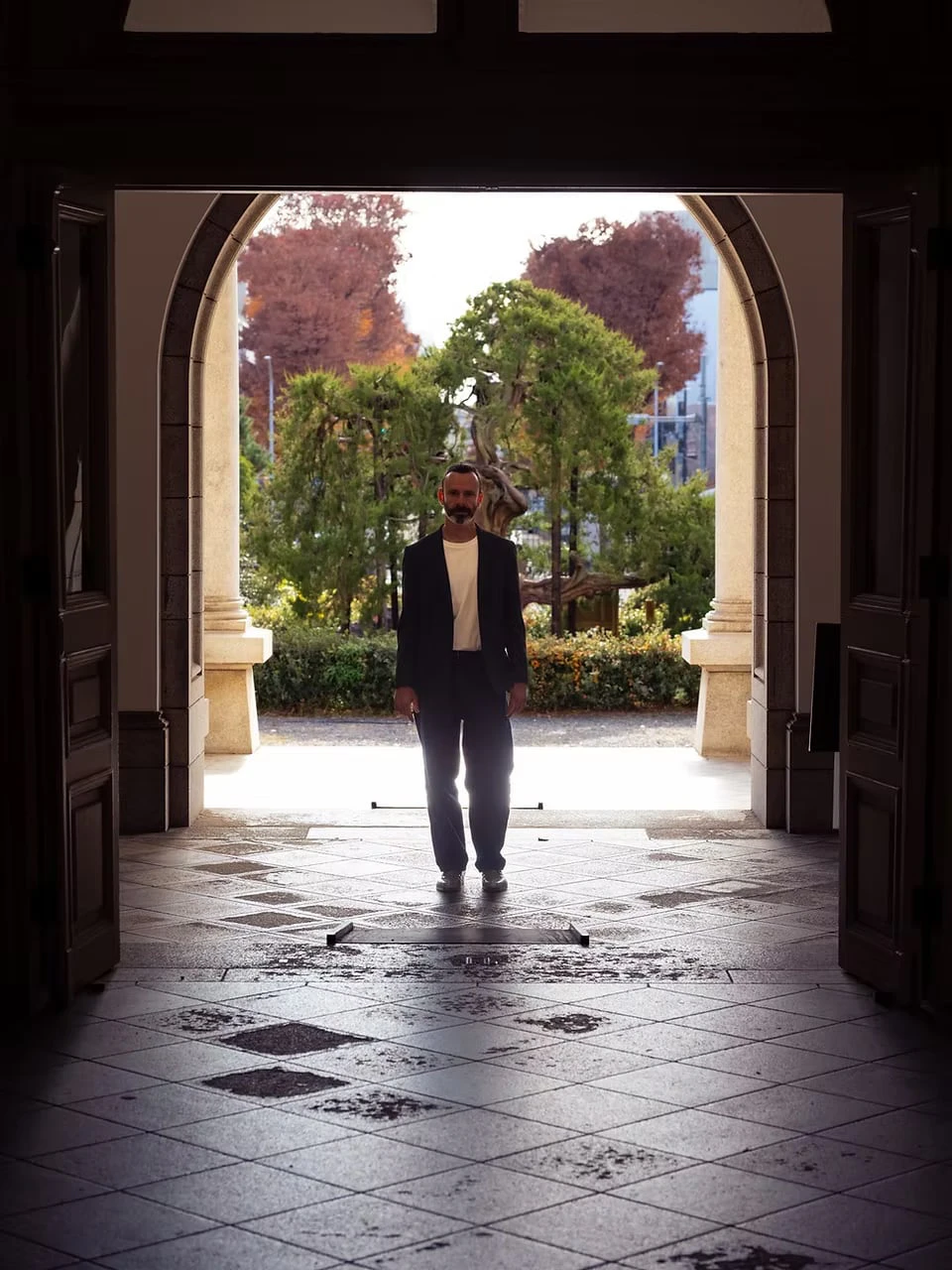 The silhouette of a man standing in a tiled entrance hall, framed by open doors and a bright garden beyond