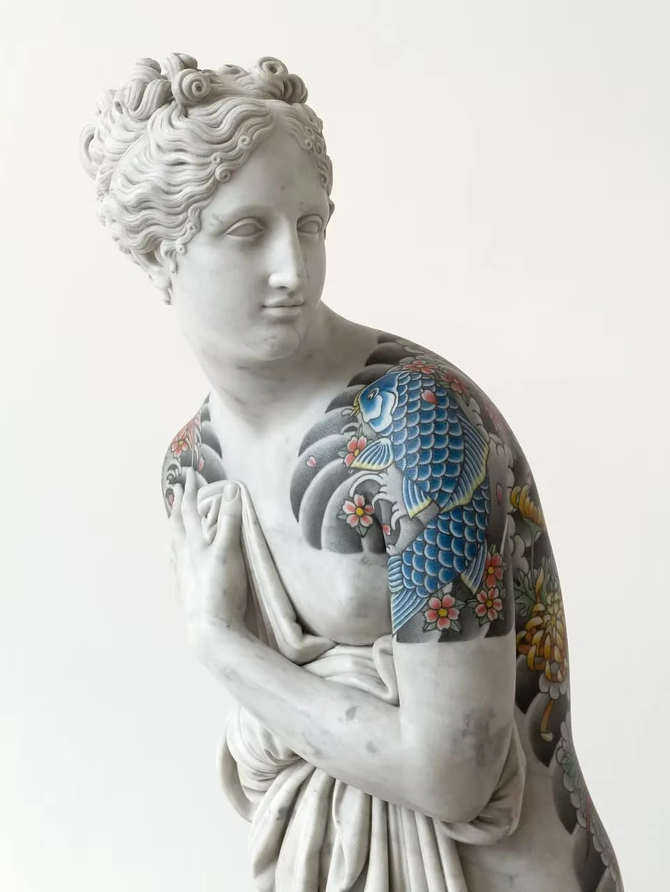 The upper body of a marble female figure against a white background, her curled hair pinned up and a cloth held to her chest, a blue carp, cherry blossoms, chrysanthemums and waves tattooed across her shoulder and arm