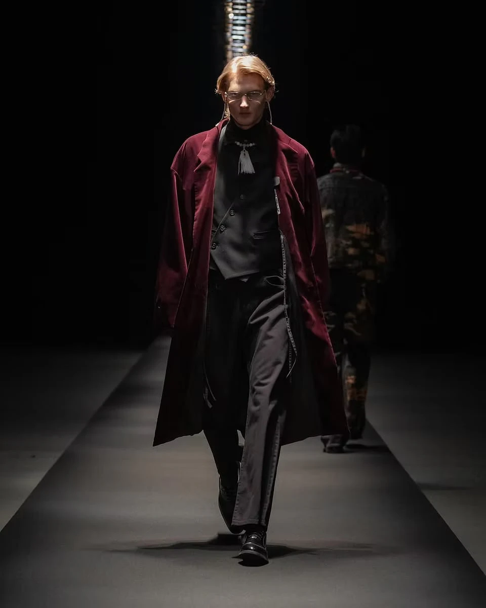 A model on the runway in a long burgundy coat over a black waistcoat and trousers, with a tasselled necklace