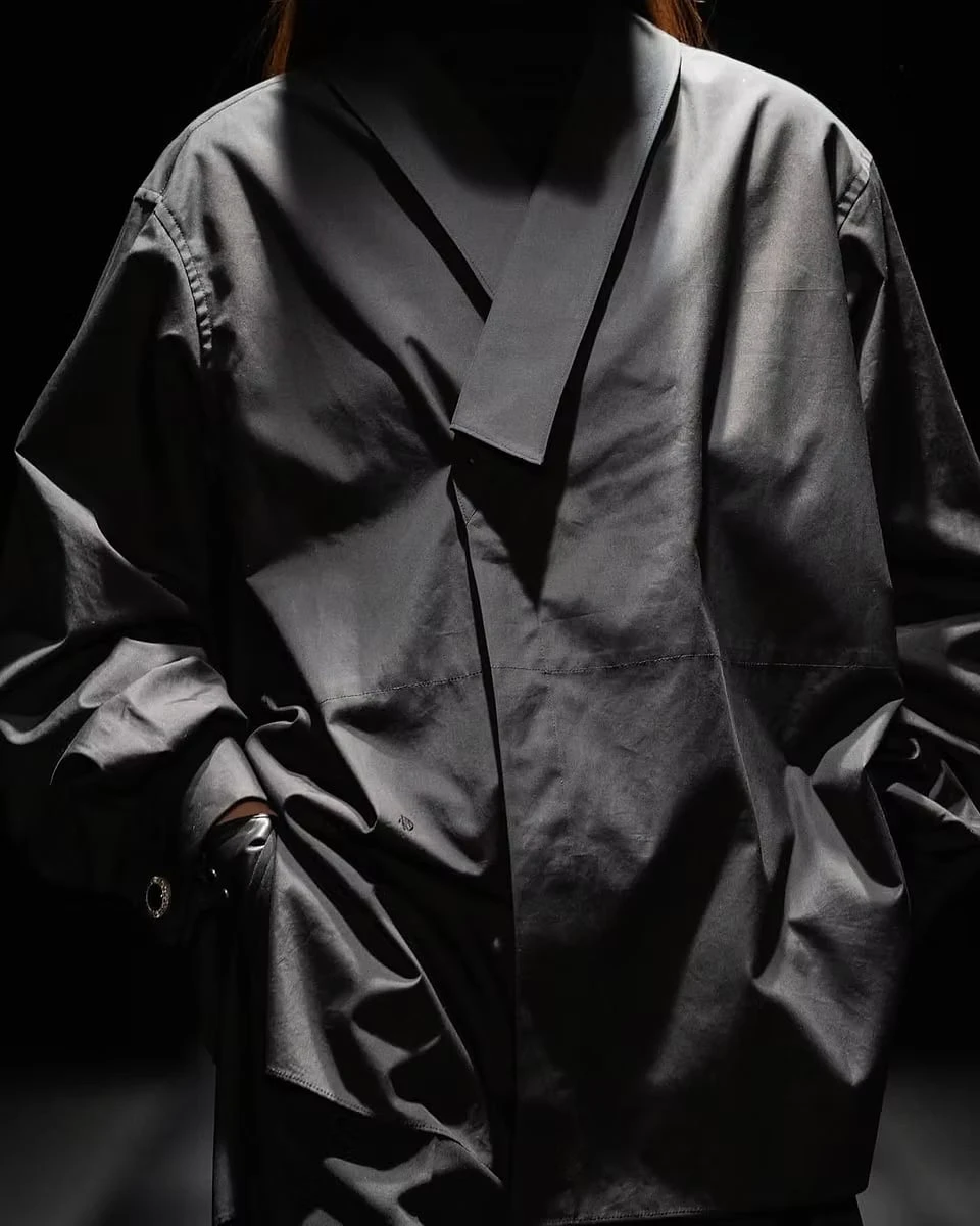 Close-up of a grey garment in low light, its diagonally overlapping collar and soft folds catching the light