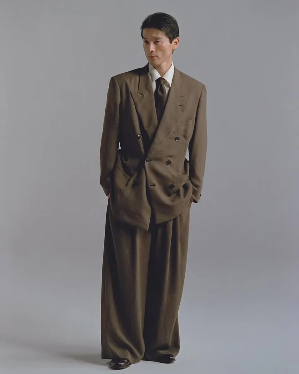 A man standing against a grey backdrop in a brown double-breasted jacket and wide, pleated trousers
