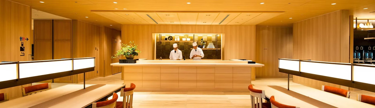 A room panelled in pale wood, with two chefs in white standing behind a long counter and rows of seats with orange chairs in the foreground