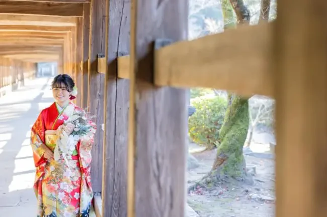 吉備津神社の回廊、木の柱越しに微笑む振袖姿の新成人女性が季節の花束を手に持つ様子