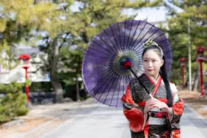 紫の和傘をさし赤い振袖姿でロケーション撮影に臨む女性が参道に立っている様子