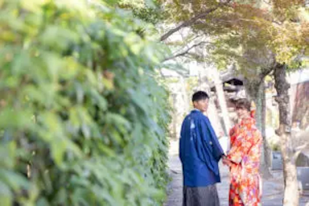 緑豊かな小道で手をつなぎ歩く紋付袴姿の新郎様と赤い着物の新婦様