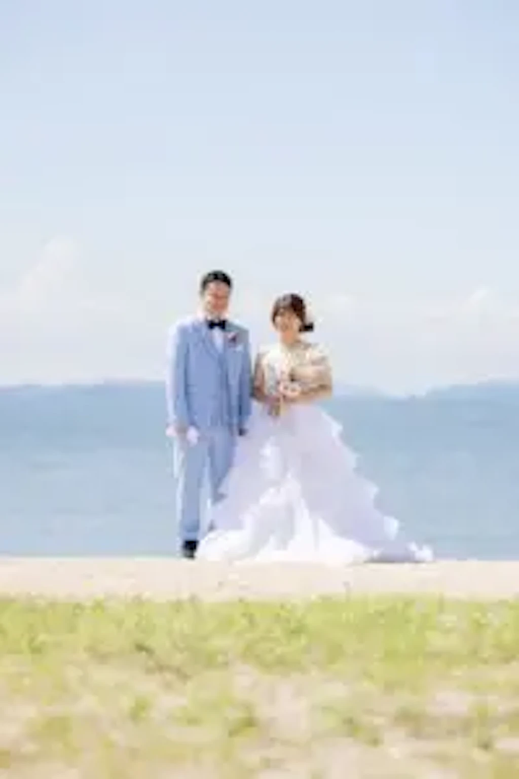 玉島沙美海岸の青空の下、白いドレスの新婦様と新郎様が寄り添って立つ全身ショットです