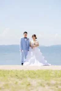 玉島沙美海岸の青空の下、白いドレスの新婦様と新郎様が寄り添って立つ全身ショットです