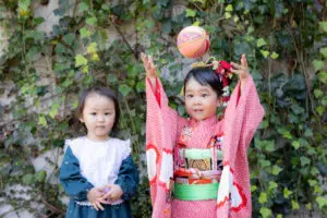 緑の蔦を背景に、着物姿の姉と私服のいもうとが並んで笑顔を見せる七五三のワンシーン