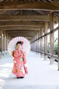 吉備津神社の長い回廊を背景に、和傘を手にピンクの着物姿で微笑む七五三の女の子